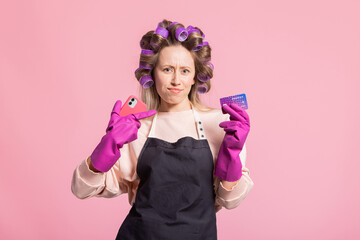 Young woman points finger at credit card, holds phone in hand, has cleaning gloves on, orders chemicals via online store app, home shopping, housewife on pink background.