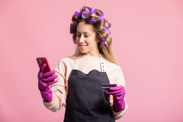 Smiling woman with rollers on head poses against pink background holds credit card in hand...