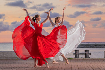 Two ballerinas in a white and red flying skirt and leotard dancing in a duet on the embankment of the ocean or sea against the backdrop of the sunset sky