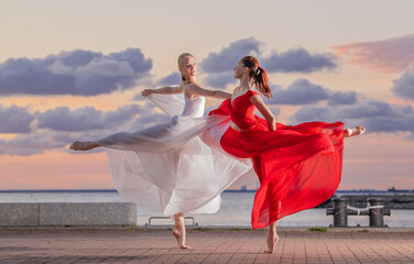 Two ballerinas in a white and red flying skirt and leotard dancing in a duet on the embankment of the ocean or sea against the backdrop of the sunset sky