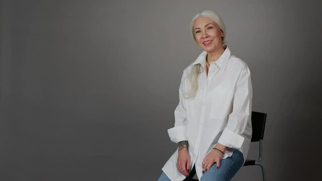 Portrait of modern grey haired woman in oversized white shirt, blue jeans and arm tattoo smiling at camera sitting on chair at dark grey studio background