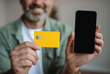 Smiling elderly european man with beard shows smartphone with blank screen and credit card in room