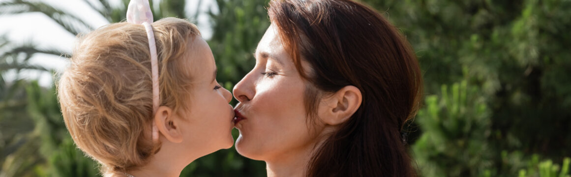Side view of brunette mother kissing baby daughter outdoors in Valencia, banner.