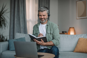 Smiling happy aged european man with beard in headphones has meeting remotely at computer