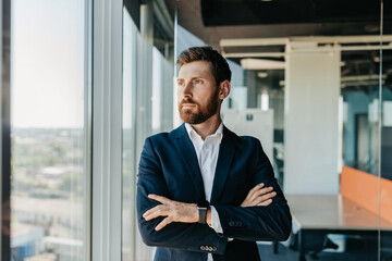 Successful businessman in formal wear standing with folded arms in modern coworking office and...