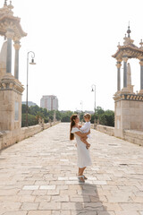 Side view of woman in white dress hugging toddler daughter on Puente Del Mar bridge in Valencia.