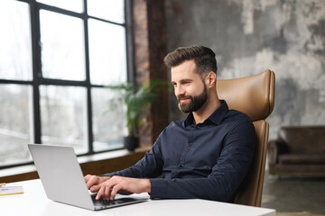 Concentrated determined young bearded office worker in business casual clothes, sitting at the desk, typing on laptop, businessman professional is working on project, writing email to client