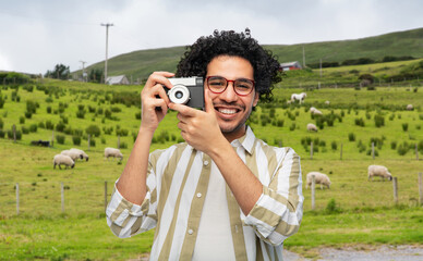 photography, travel and tourism concept - happy smiling tourist man or photographer in glasses with film camera over sheep grazing on field of connemara in ireland background