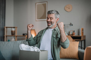 Satisfied glad excited old european male blogger with beard unpacks box and rejoices to new sneakers, makes win gesture