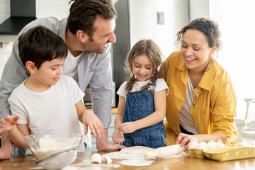 Happy family of four cooking in the kitchen together, parents and two kids making dough together, preparing pie or cookie, have a fun on weekend
