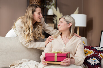 Loving woman adult daughter congratulating mature mother, hugging, handing red ribbon box with...