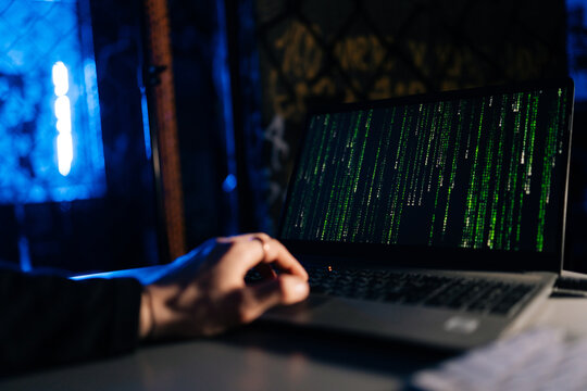 Close-up of unrecognizable hacker engaged in hacking into security system on laptop green binary code on screen with sitting at desk in dark basement room, with blue neon lights.