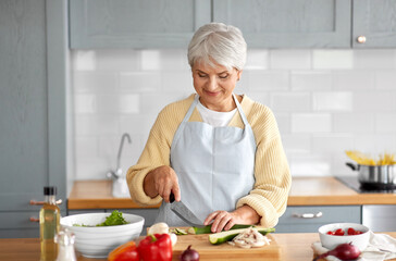 healthy eating, food cooking and culinary concept - happy smiling senior woman with knife chopping zucchini on kitchen at home