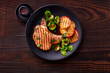 gegrilltes Kotelett mit Rosenkohl und gegrillten Äpfeln auf schwarzem Teller und Holzfond mit Barbecue Sauce