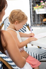 Side view of toddler child drawing on notebook near mom in outdoor cafe.