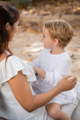 Side view of woman in white dress holding baby daughter in blurred park.