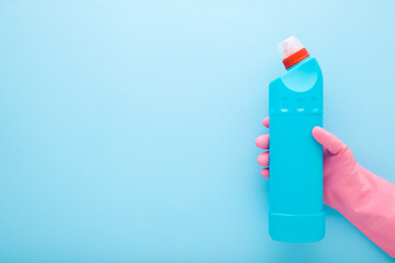 Hand in pink rubber protective glove holding toilet cleaner bottle on light blue wall background....