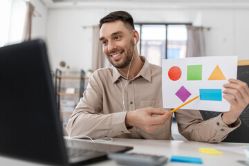 distance education, primary school and remote job concept - happy smiling male teacher with laptop and picture of geometric shapes in different colors having online class at home office