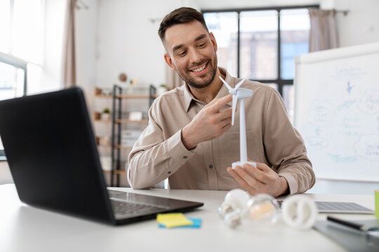 distance education, school and green energy concept - happy smiling male teacher with laptop computer and wind turbine model having online class at home office