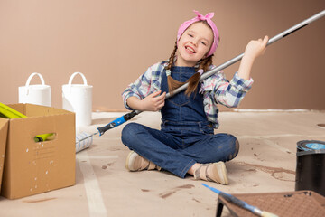 Smiling girl sits on the floor with legs crossed holds a paint roller on a long stick in front of a tray of brown colored paint, help in renovation painting walls.