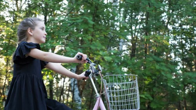 Teenager girl wearing black dress rides bike with basket at back sunlight. Stylish girl rides bicycle with excitement and happy expression past park trees
