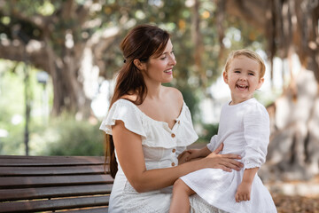 Brunette woman looking at daughter laughing on bench in park.