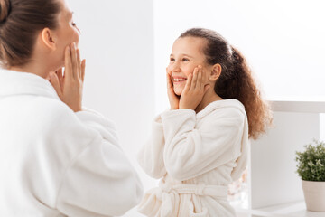 beauty, hygiene, morning and people concept - happy smiling mother and daughter touching their faces in bathroom