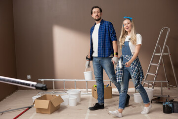 Young couple in love walks into the living room of a new apartment during renovations. The man carries a can of paint in hand, the woman holds a wall roller, renewing the color of the room.