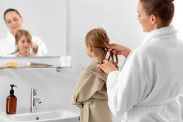 beauty, family and people concept - happy smiling mother and daughter looking to mirror and braiding hair in bathroom