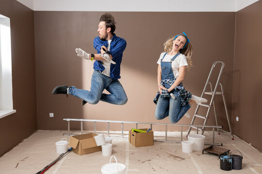 Photo Of Young Couple In Love In A Hop. Happy Woman With Man Having Fun During Renovation, Painting Apartment, End Of Finishing Work Joy Jumping Up In Background Ladder Paint Rollers Cans.