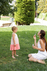 Side view of woman blowing soap bubbles near child in dress in park.