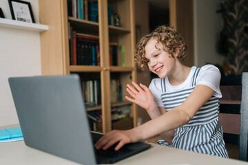 Portrait of cheerful child girl sitting at table having video call, looking at laptop display and waving hand to friends, family, parents, teacher. Preteen girl bed making videocall via laptop at home