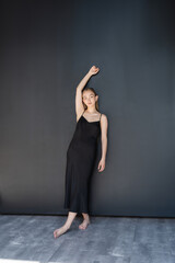 full length of barefoot woman in black strap dress posing with raised hand near dark wall.