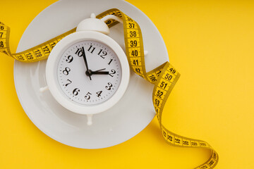Diet concept. Alarm clock on a white plate with measuring tape on a yellow background