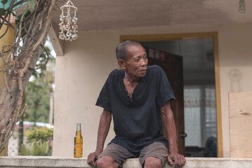 A poor middle aged man relaxes and bums around while sitting on the ledge of the house veranda. An...