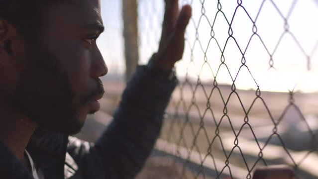 Male prisoner looking through a wire fence thinking about life and showing regret about bad choices. Young african man worried about future behind bars outside in a prison yard after doing crime