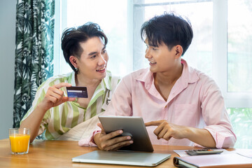 Obraz premium Cute Asian LGBT gay man couple, using tablet online shopping, holding credit card, spend time together. Healthy lifestyle with orange juice.