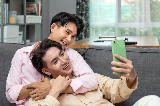 Cute Happy Smiling Asian LGBT Homosexual Men Or Gay Couple Sitting And Reclining On Sofa, Embracing And Selfie Photo Together. Looking At Camera.