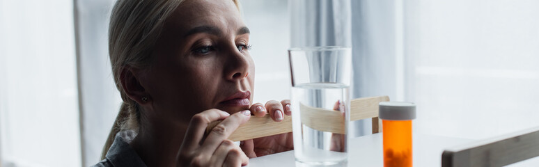 blonde woman with menopause looking at medication and glass of water on table, banner.