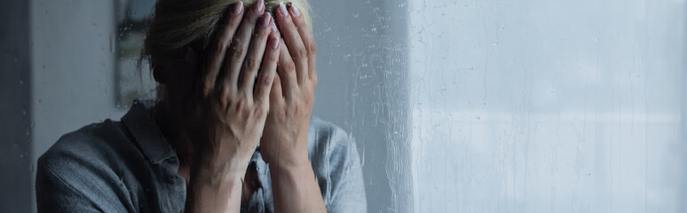 depressed blonde woman covering face behind wet window with rain drops, banner.