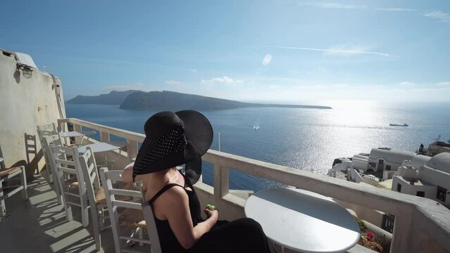Black dressed elegant woman enjoy view of Oia on Santorini island sitting on traditional taverna with old wooden table and chairs balcony