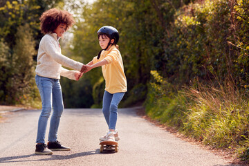 Boy Teaching Girl To Ride Skateboard Holding Her Hands On Country Road