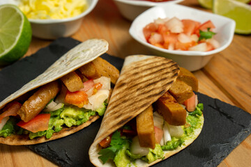 45º view of two vegan guacamole and seitan tacos served on a blackboard with lime, cheese and pico de gallo, on a wooden table with a red cloth behind. Horizontal image