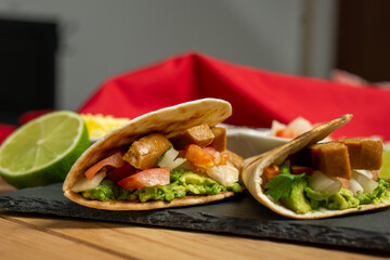 Side view of two vegan guacamole and seitan tacos served on a blackboard with lime and pico de gallo, on a wooden table with a red cloth behind. Horizontal image