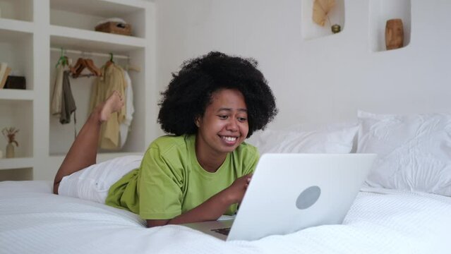 A mixed race woman lies on a bed in a hotel and communicates with relatives via video link on a laptop from his bedroom. A black woman with an afra haircut uses the internet on a laptop.