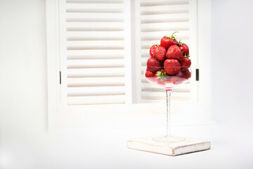 Strawberries in a tall elegant wine glass. A white window with shutters in the background. Summer season. Space for copying.