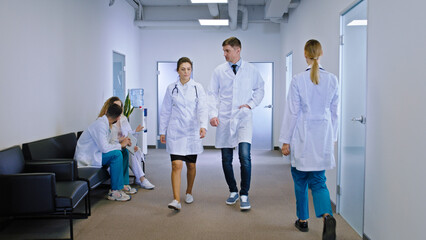 Female and man doctors walking through the modern corridor in the hospital some other nurses and doctor on the chair have a discussing as well