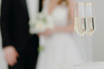 Two glasses of champagne stand on the table against the background of newlyweds in love at the wedding ceremony.