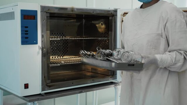 Female nurse in white protective suit taking clean sterilized surgical tools and instruments from medical autoclave steam sterilizer machine, healthcare worker in hospital operating room, slow motion.