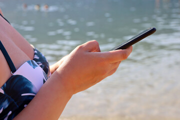 Woman in bikini with smartphone on a beach, mobile phone in female hands on sea background. Concept of using phone in travel, sms, online communication at the seaside